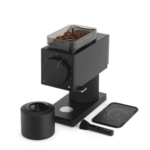 The image displays electric grinder Ode Gen 2 by Fellow. The electric grinder is black. In the image it is also possible to check the coffee dispenser, the cleaner accessory and coffee beans inside the electric grinder.