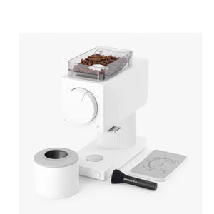 The image displays electric grinder Ode Gen 2 by Fellow. The electric grinder is white. In the image it is also possible to check the coffee dispenser, the cleaner accessory and coffee beans inside the electric grinder.