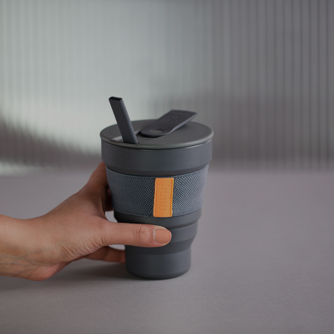 The image displays a hand holding a reusable cup in the colour grey from the brand Hunu. The reusable cup have an orange detail and both wall and floor are greay.